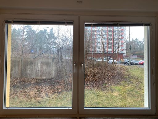 Fönster med suddig utsikt över en grön gräsmatta och en bygård i bakgrunden.